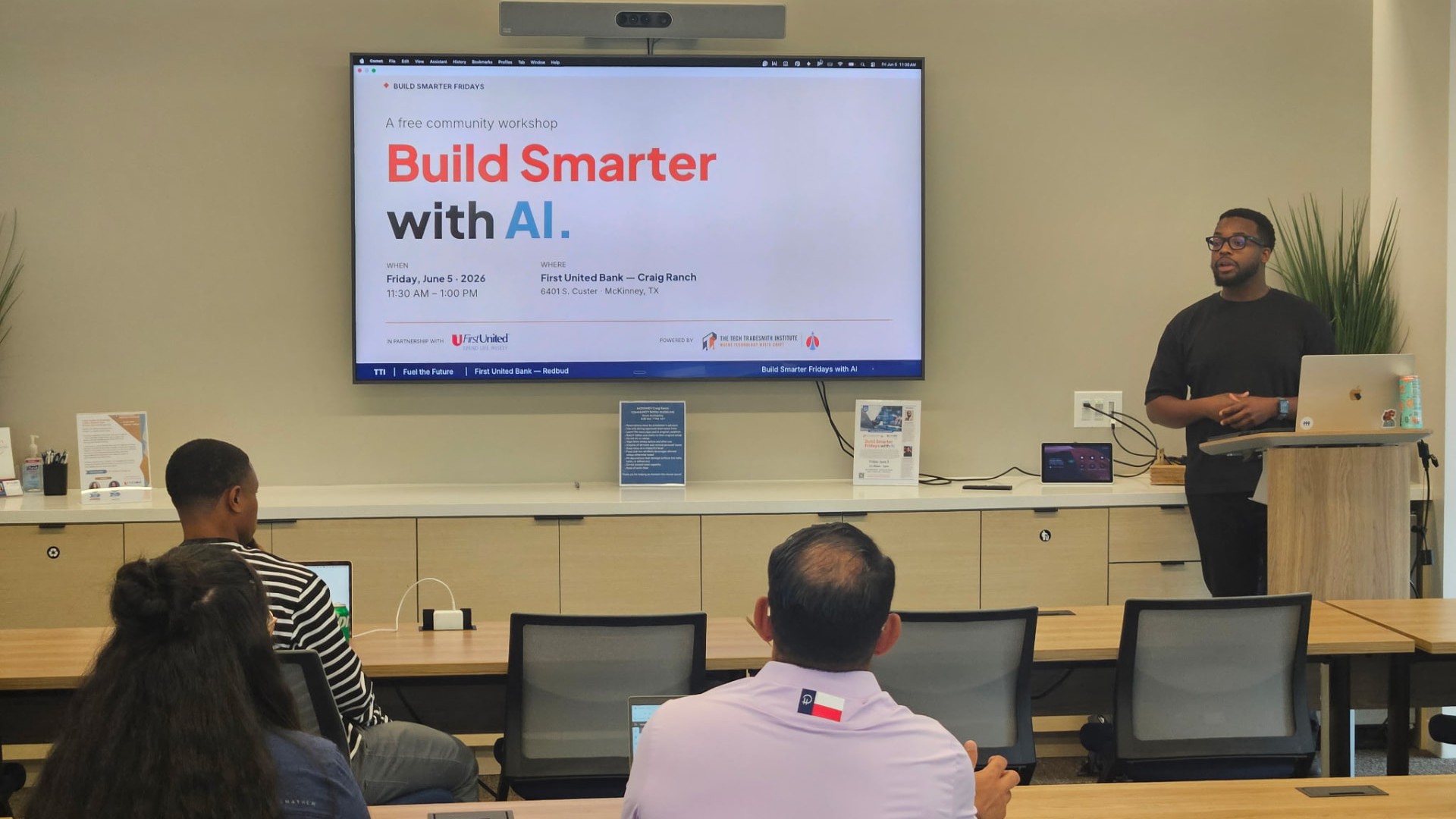 A Build Smarter Fridays session in progress: a presenter at a lectern speaking to a small group seated at tables, with the session slide on the wall screen behind him.